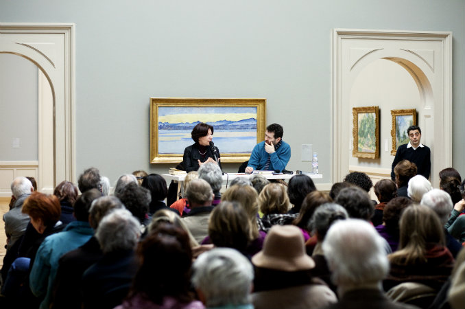 Deux conférenciers - Sylviane Dupuis et Pierre Lepori - s'adressent à un public nombreux devant un tableau de Ferdinand Hodler.