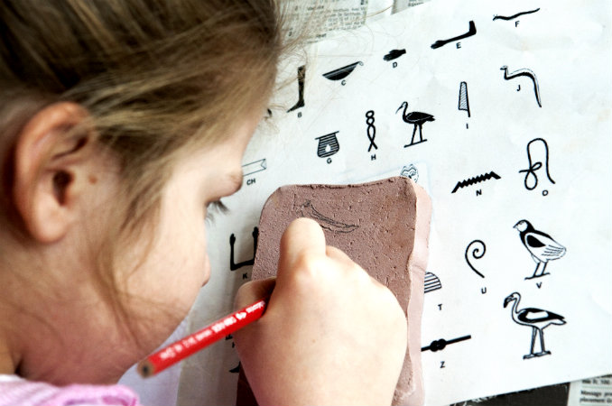 Une petite fille écrit sur une tablette de terre en s'aidant d'une table d'équivalence entre les hiéroglyphes égyptiens et l'alphabet latin